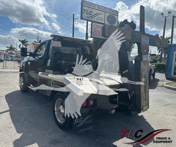 Tow truck with crane arm and rear hitch, TLC Truck & Equipment logo, visible signs in background, truck bed and wheel. Text on truck includes TLC branding and contact area.