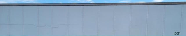 Image shows a light gray metal wall of a building with a thin blue sky edge; no visible text other than a small 53' marking on the lower right corner.