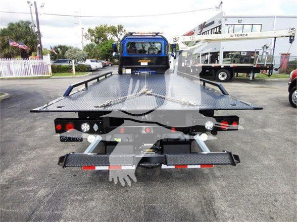 Flatbed tow truck bed with hydraulic ramps and rear lights visible, Peterbilt 337 in background, outdoor dealership lot.