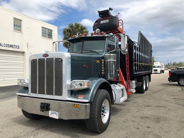 Peterbilt 388 dump truck with grapple attachment, front grille, large chrome bumper, two chrome exhaust stacks, Serco grapple unit on top, green cab, black dump bed, side windows, and road in background.