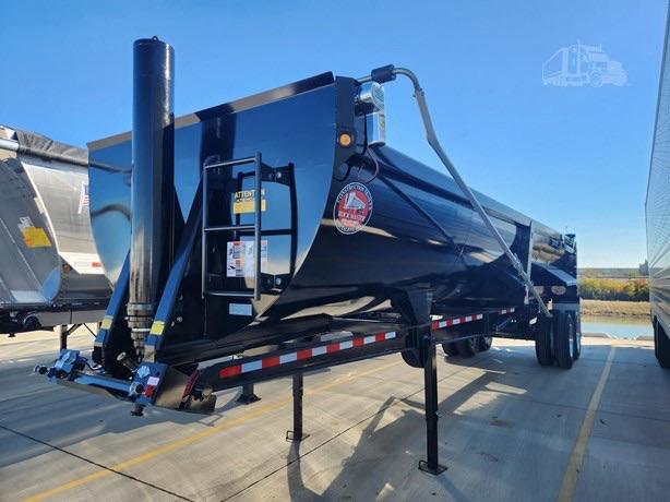 End-dump trailer with blue paint, steel end dump body, front ladder and support legs. Visible labels on side include a round red/white sticker and safety placards.