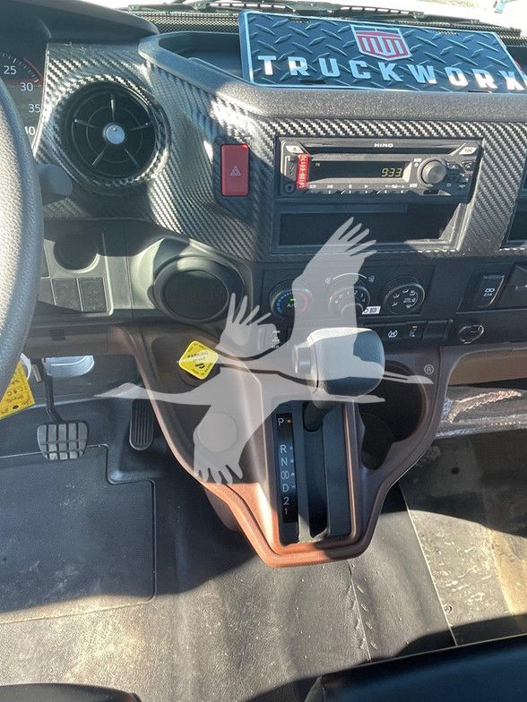 Cab interior of a 2025 Hino L6 showing center console with Hino stereo, climate controls, and automatic gear selector. Dashboard features carbon-fiber style trim and V-shaped gear shifter with PRND2 text visible.