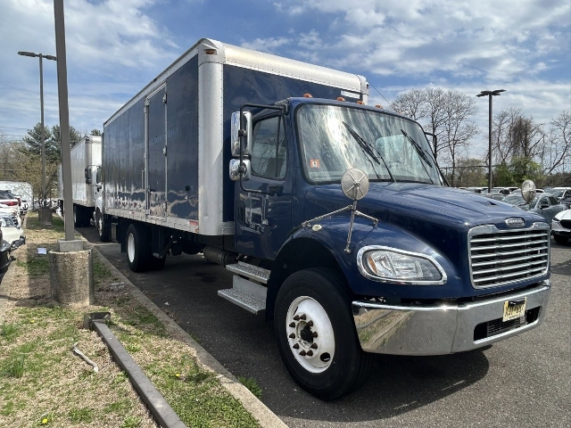 Freightliner M2 106 box truck in blue with silver cargo box, side steps, large side mirrors. Front grille chrome, round headlights, front bumper chrome, white wheels. Vehicle shows stock number marking and VIN citation in listing.