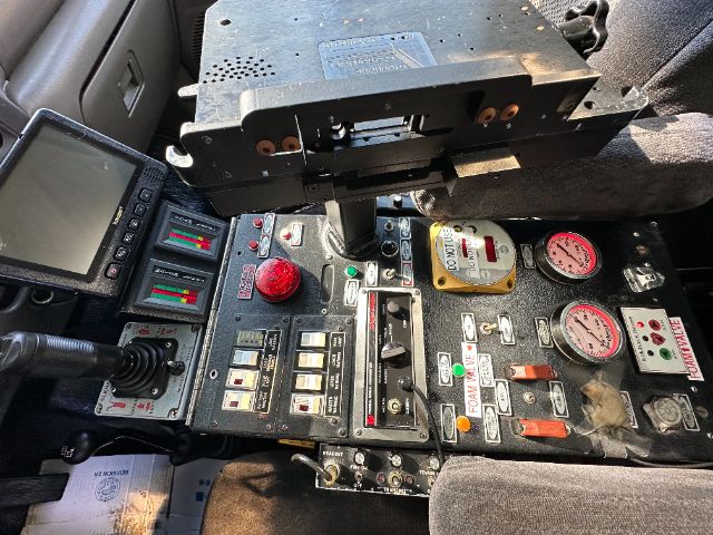 Dashboard of a Ford F-550 fire truck cab showing control panel with gauges, switches, and a red emergency stop button. A monitor is on the left and a metal rack above the panel.