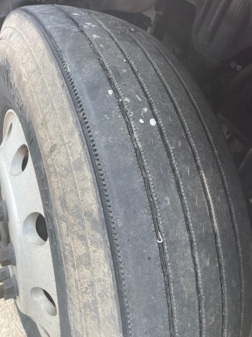 Truck tire close-up showing tread wear on highway steered axle, with metallic wheel rim beside.