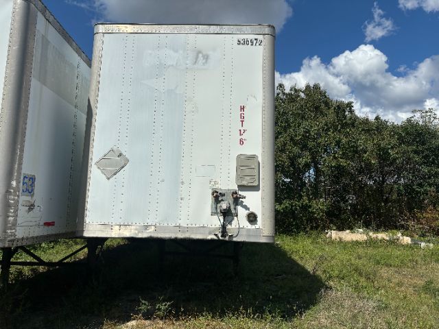 Rear view of a white Wabash trailer with vertical dimensions marked 8'6" and a hitch/docking area, showing a road/grass field and blue sky.