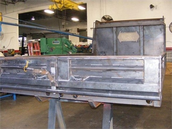 Dump body truck bed on a metal stand in a workshop, showing heavy-gauge aluminum panels and rear tailgate with chain latches.