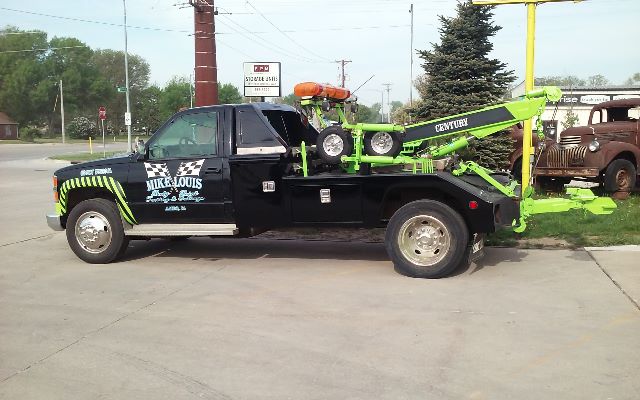Tow truck with a green Century tow boom mounted in the rear, black cab with Missouri label, orange beacon on cab roof, and spare wheel on flatbed.
