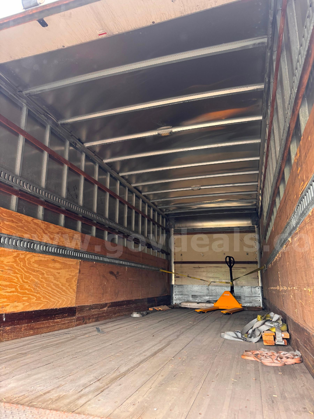 Inside Freightliner M2 106 box truck cargo area showing wood panel walls, metal rails, and loading equipment including a pallet jack and straps.