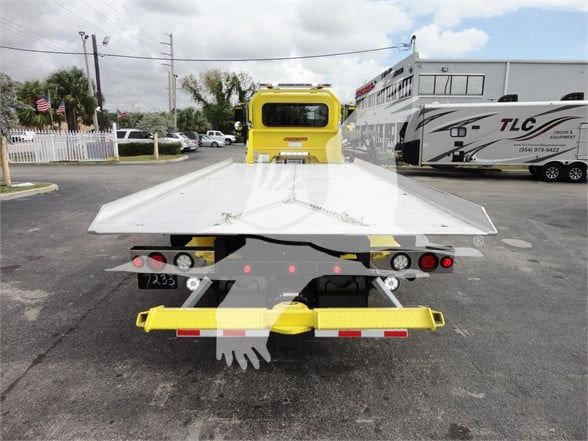 Rear view of a yellow Peterbilt 337 towing truck with a flatbed ramp, license plate area, lights, and a secondary vehicle in background.