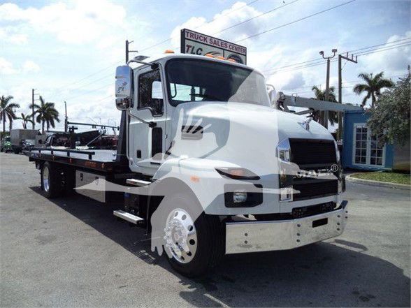 2024 Mack MD6 tow truck in a dealership lot, white cab with flatbed, front bumper, large side mirrors, and tires visible.