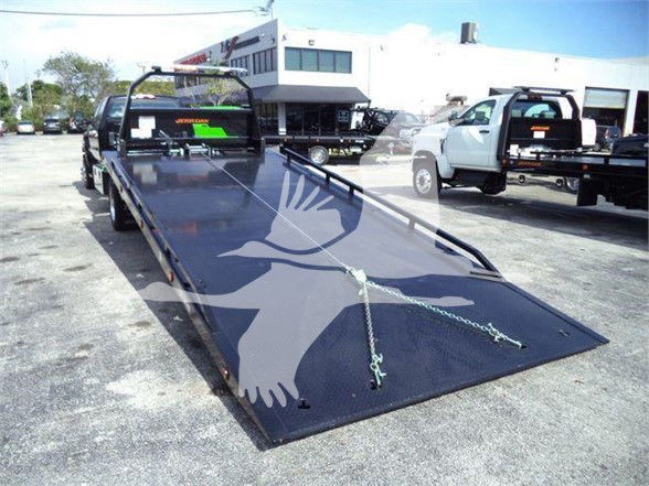 Flatbed tow truck with a long black steel ramp and white pickups in background. Visible text on the truck: looks like a logo or markings on the cab area.