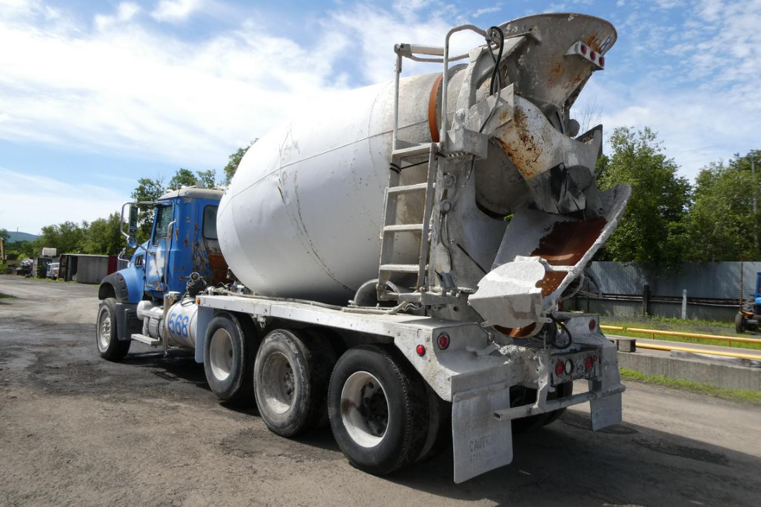 Concrete mixer truck with a blue cab and large white drum, rear chute and ladder, showing triple rear axles and worn metal surfaces.