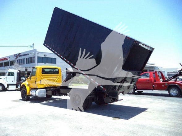 Truck with a raised dump bed showing a yellow International truck and other equipment in the background.