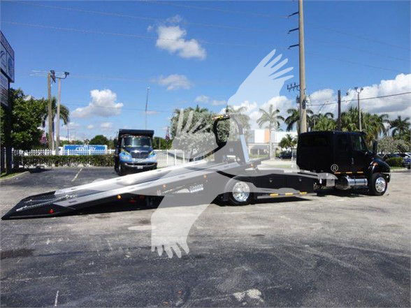 Tow truck with a lowboy trailer attached, in a parking lot. Visible text on the vehicle: none clearly legible due to watermark. The background shows a blue sky with clouds and palm trees.