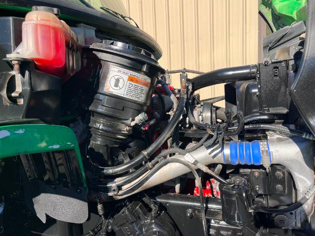 Truck engine bay showing various components including a black circular reservoir with warning label, red coolant reservoir, blue hose clamp, and various hoses and metal piping.