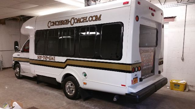 Rear and side view of a white Ford E-450 bus with black windows and brown/black stripe; text on bus includes 'Concorde Coach' and contact numbers; rear has a tailgate.