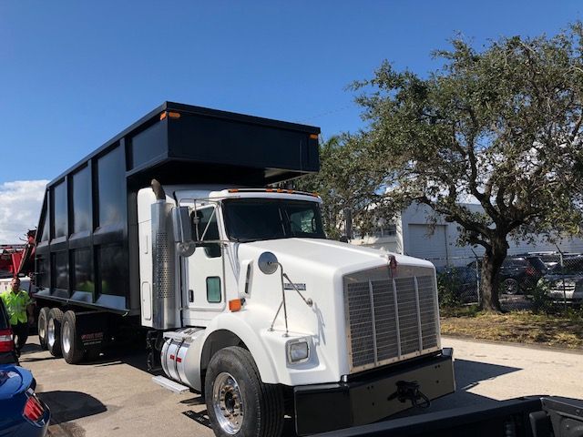 Kenworth T800 dump truck with a large black dump bed; white cab; front grille; side mirrors; dual rear axles visible; clear sky.