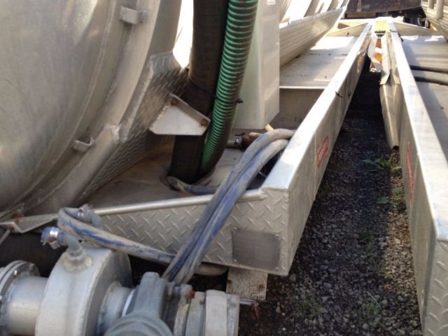 View of a vacuum tank truck chassis with metal diamond plate steps, hoses, and piping. Visible are a green corrugated hose, a gray power line, and a cylindrical tank along the left side.