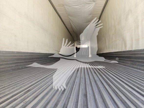 Interior of a refrigerated trailer with ribbed metal floor, long white walls, and an opening at the far end; watermark of a flying bird placed over the image.