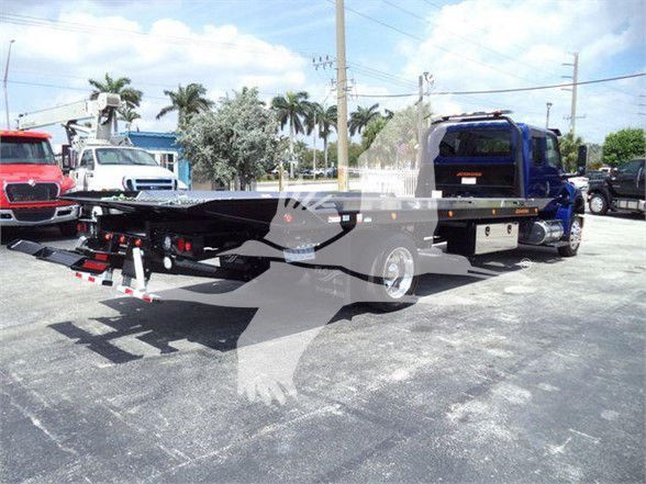 Tow truck chassis with flatbed on International MV, blue cab, parked lot with other trucks in background.