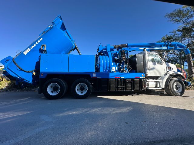 Blue Sterling L7500 vacuum truck with loader crane, side view. Text on blue hopper reads 'macon' or similar branding. Vehicle has chrome cab, multiple axles, hoses and equipment.