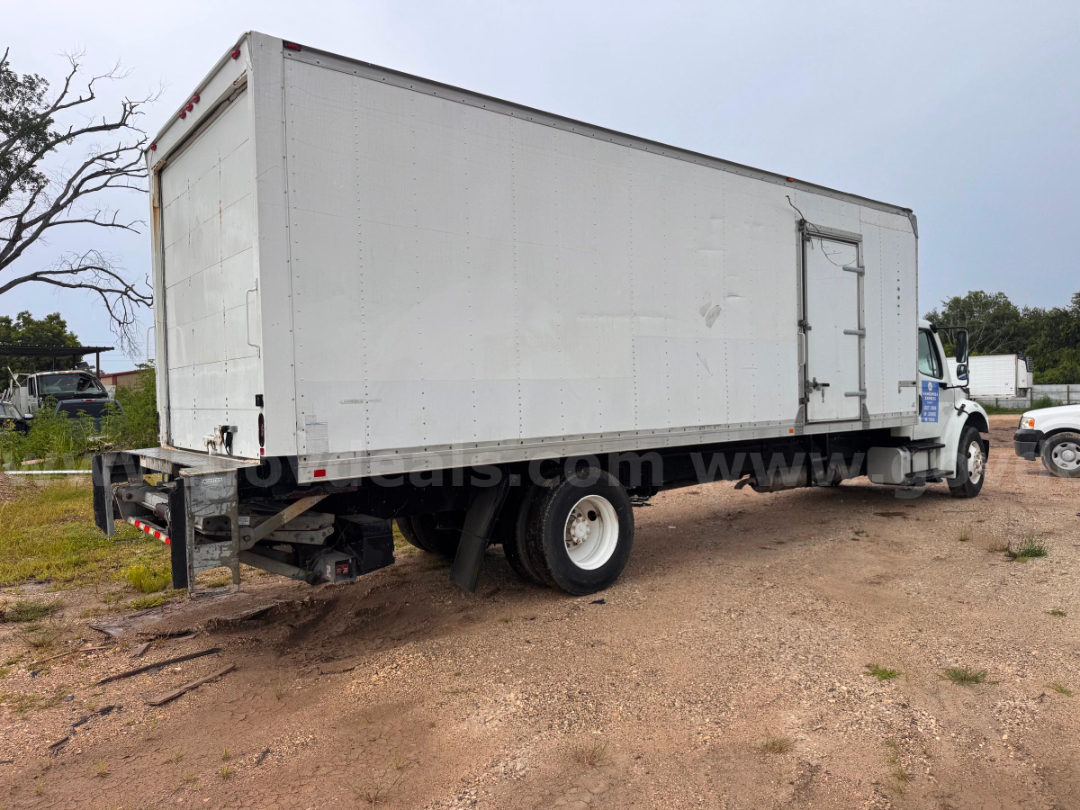Box truck in a dirt lot showing a white Freightliner M2 106 box body with rear underrun guard and left-side cab door. Visible: stock number plate, VIN context, and dual rear wheels.