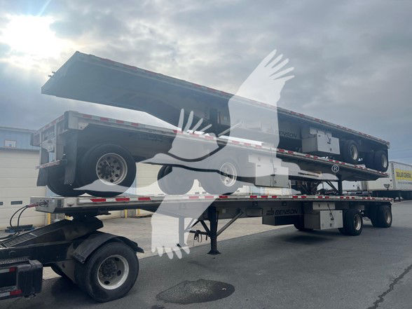 Flatbed trailer loaded on a two-tier carrier being transported, showing a Benson aluminum flatbed with wheels and support stands.
