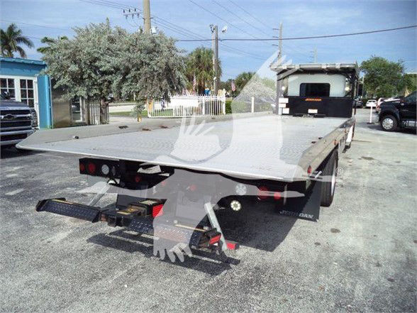 Flatbed tow truck bed with rear bumper, taillights, and equipment resembling a wheel lift. Visible on deck: no specific markings legible. Tow truck chassis visible in distance.