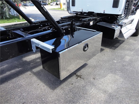 Kenworth W900 chassis showing a black rectangular toolbox mounted under the cab with a circular keyhole, part of the truck frame and fuel/tool compartments visible.
