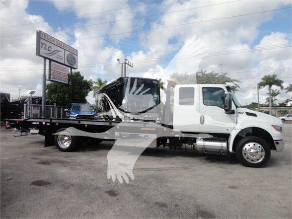 2025 International MV tow truck in a dealership lot showing a white cab and flatbed with a black crane/haul bed. Visible text on signboard in background and on truck not legible.