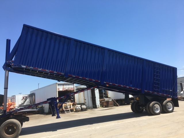Trailer in-service blue dump trailer with raised bed and tandem axle, side view showing loading ramp and plumbing.