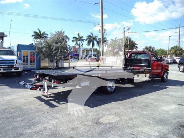Flatbed tow truck with a red cab in a parking lot, Chevrolet Silverado 6500HD listing visible in the background with palm trees.
