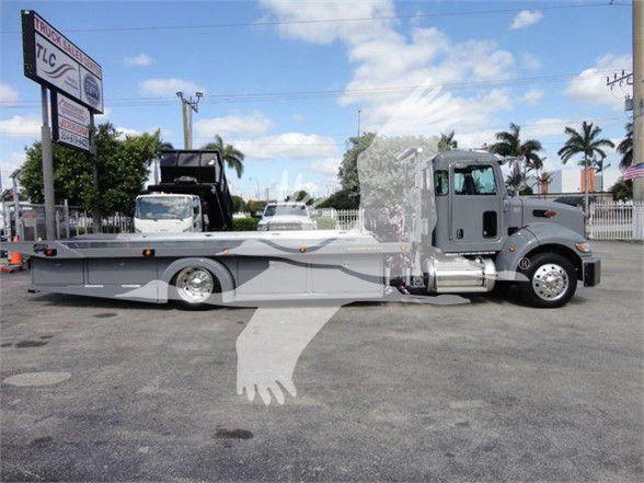 Peterbilt 337 tow truck, side view, long low-bed trailer with gray paint and graphic stripe. cab area visible with chrome fuel tanks and mirrors.