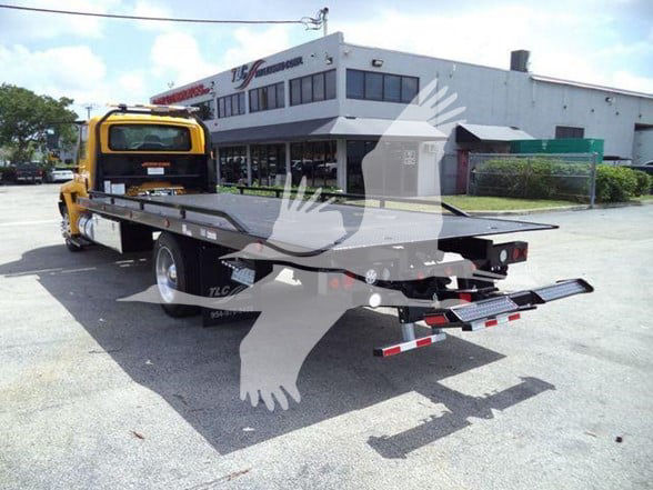 Flatbed tow truck with yellow cab and long flat steel bed, rear underride bumper, in a dealership lot.