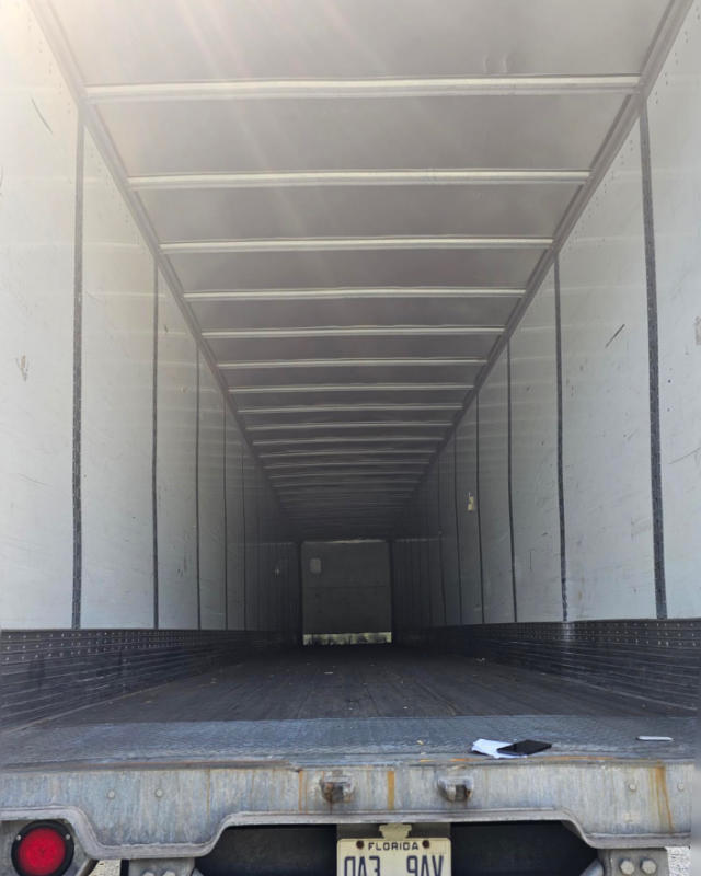 Interior of a cargo trailer with vertical ribbed walls, metal floor, and an open rear showing depth into the trailer.