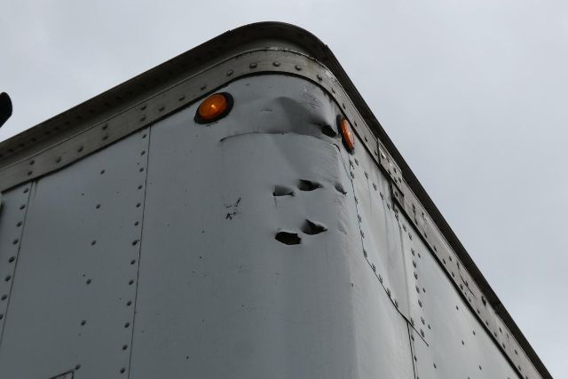 Corner of a gray Great Dane trailer with riveted metal panels, small dents and a rounded top. Round orange marker lights near the edge.