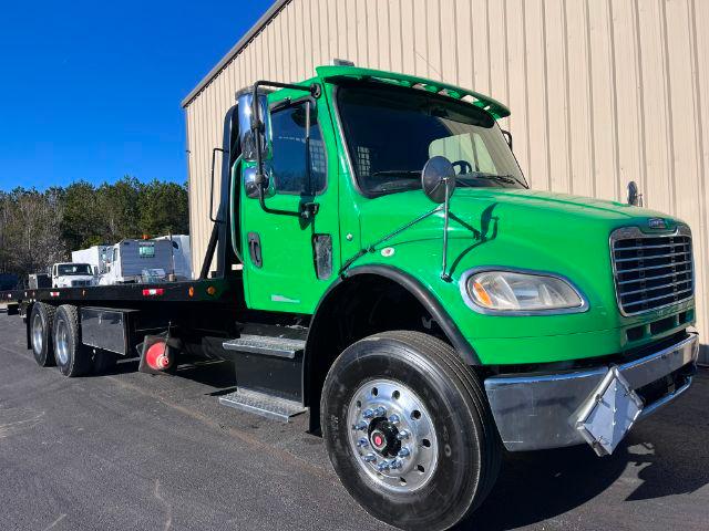 Green Freightliner M2 106 rollback tow truck cab with flatbed at a dealership, side mirror, front grille, and chrome bumper visible.
