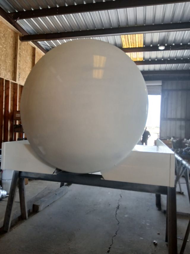 A large white spherical vacuum tank mounted on a trailer frame inside a workshop. The sphere sits on a white rectangular cradle with metal supports.