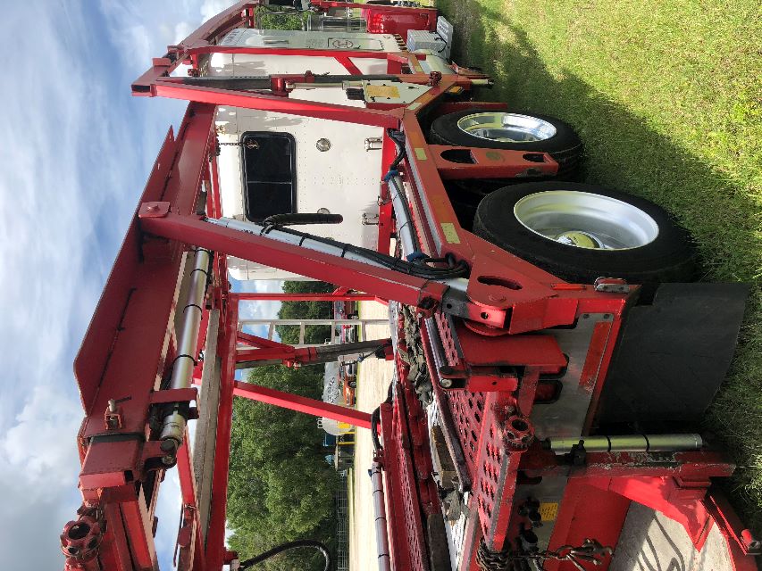 Red auto carrier with chain racks and hydraulic arms on a Peterbilt truck, side view showing large tires and white cab in background.