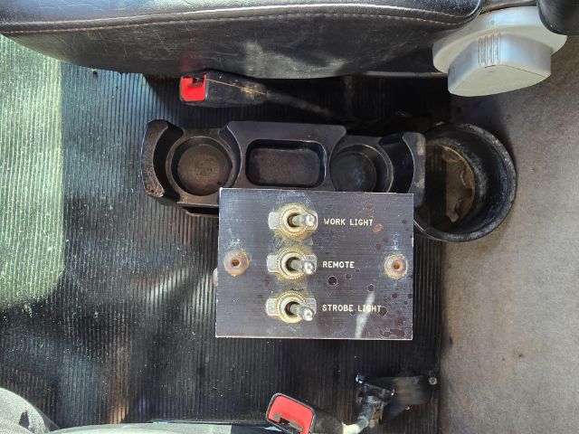 Panel with three labeled switches mounted on floor area between seats. Text reads: 'WORK LIGHT' at top, 'REMOTE' middle, 'STROBE LIGHT' bottom. Each label is next to a screw terminal. Surrounding area shows cup holders and seat rails.
