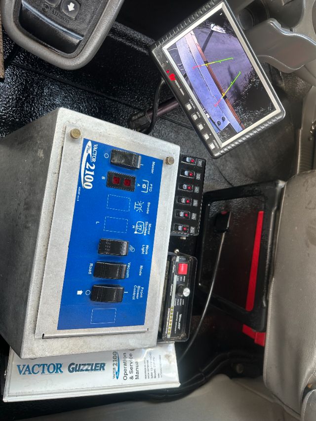 Trucks interior with a blue Vactor 2100 control panel, monitor displaying rear camera guidelines, and a Vactor Guzzler operation manual under the panel.