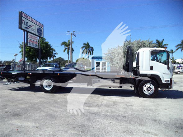 Isuzu FTR tow truck with flatbed, side view showing cab and long flatbed, dealership lot in background.