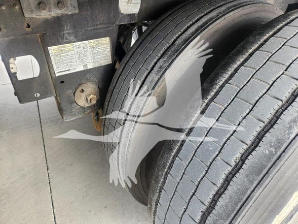 Rear wheel and chassis area of a trailer showing the suspension and tire tread. A small metal plate with warnings/labels is visible on the left side near the frame.