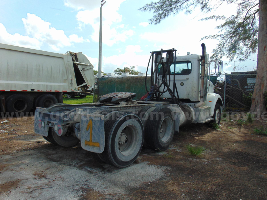 Vehicle rear and cab of a white Peterbilt 365 cab and chassis with dual rear axles, exposed fifth wheel, and hoses. Muddy ground and another truck in background.