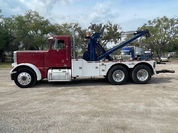 Tow truck chassis with a red cab and white service body, extending a blue hydraulic boom arm on a flatbed, 3-axle arrangement.