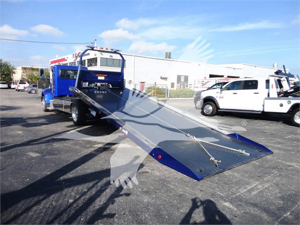 Tow truck with a long metal ramp extending from the back, blue cab, white flatbed; background shows a dealership lot with other vehicles. Visible text on the truck is not legible.