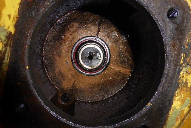 Circular mechanical component with a metal bearing inside a dark, rust-stained housing. Inner ring shows a silver center with a small hole and red seal. Surrounding area yellow-painted.