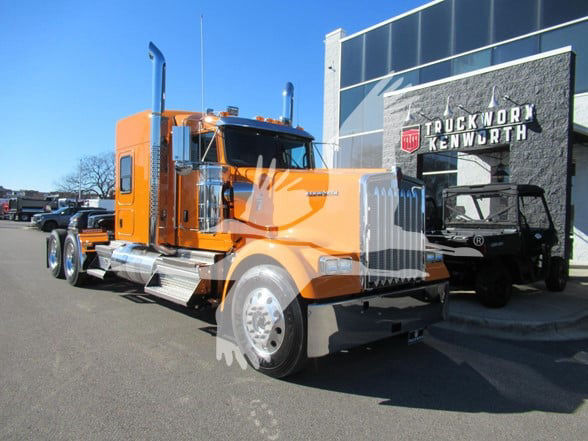 Kenworth W900L conventional sleeper truck in orange, side view at dealership with chrome exhaust stacks and large grille.