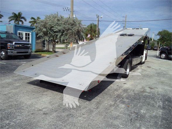 Kenworth T370 tow truck with a lowered flatbed and side ramps. Visible text: none on the bed. The truck is in a lot with other vehicles and palm trees in the background.